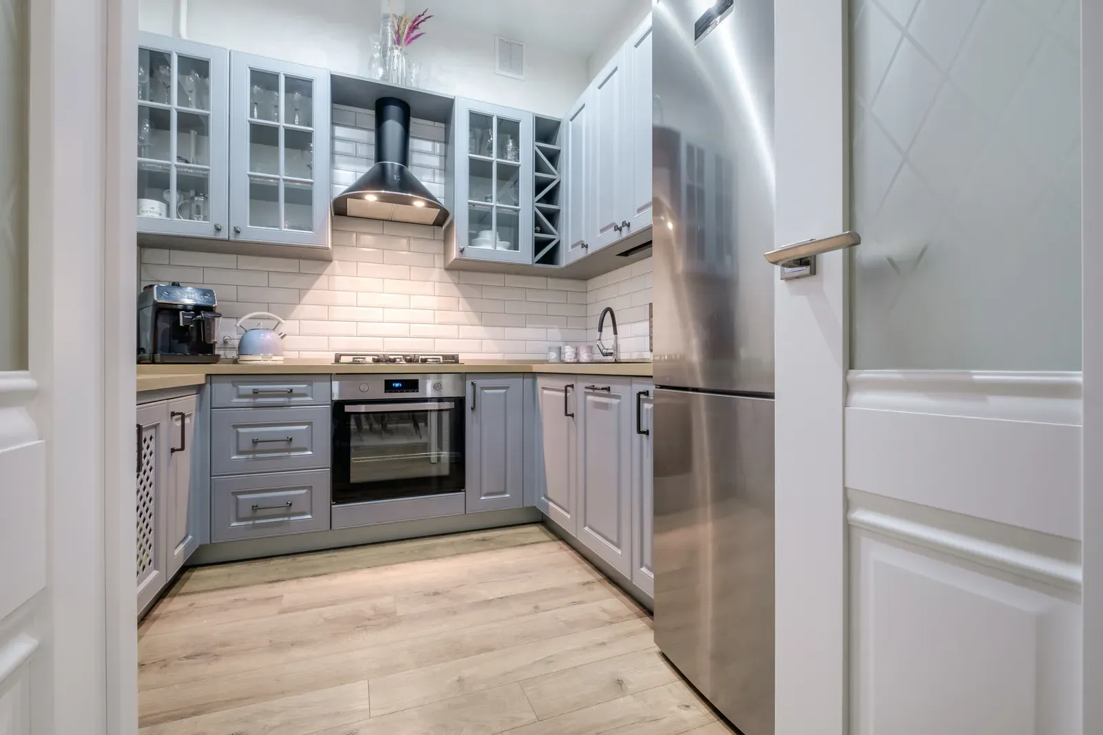 A small U-shaped kitchen in pale grey raised-panel cabinets with glass-front upper doors, a wine rack and a black range hood