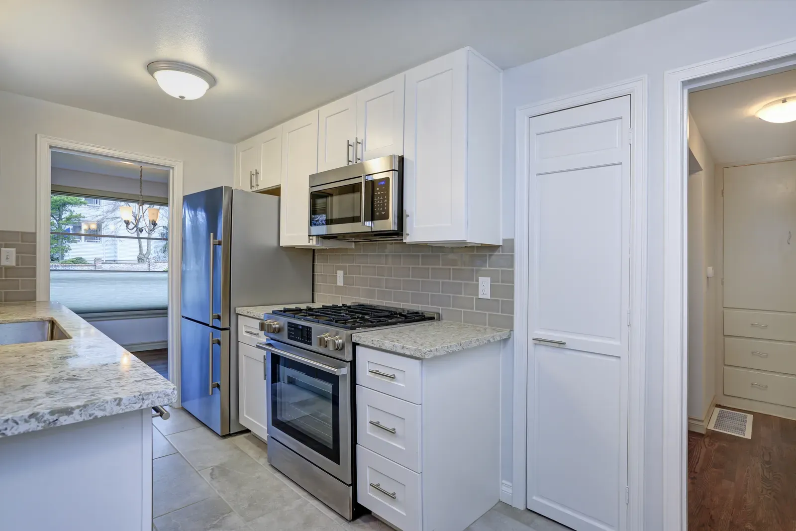 A small white shaker kitchen with a stainless gas range, a gray subway-tile backsplash and a tall pantry door beside the cabinets