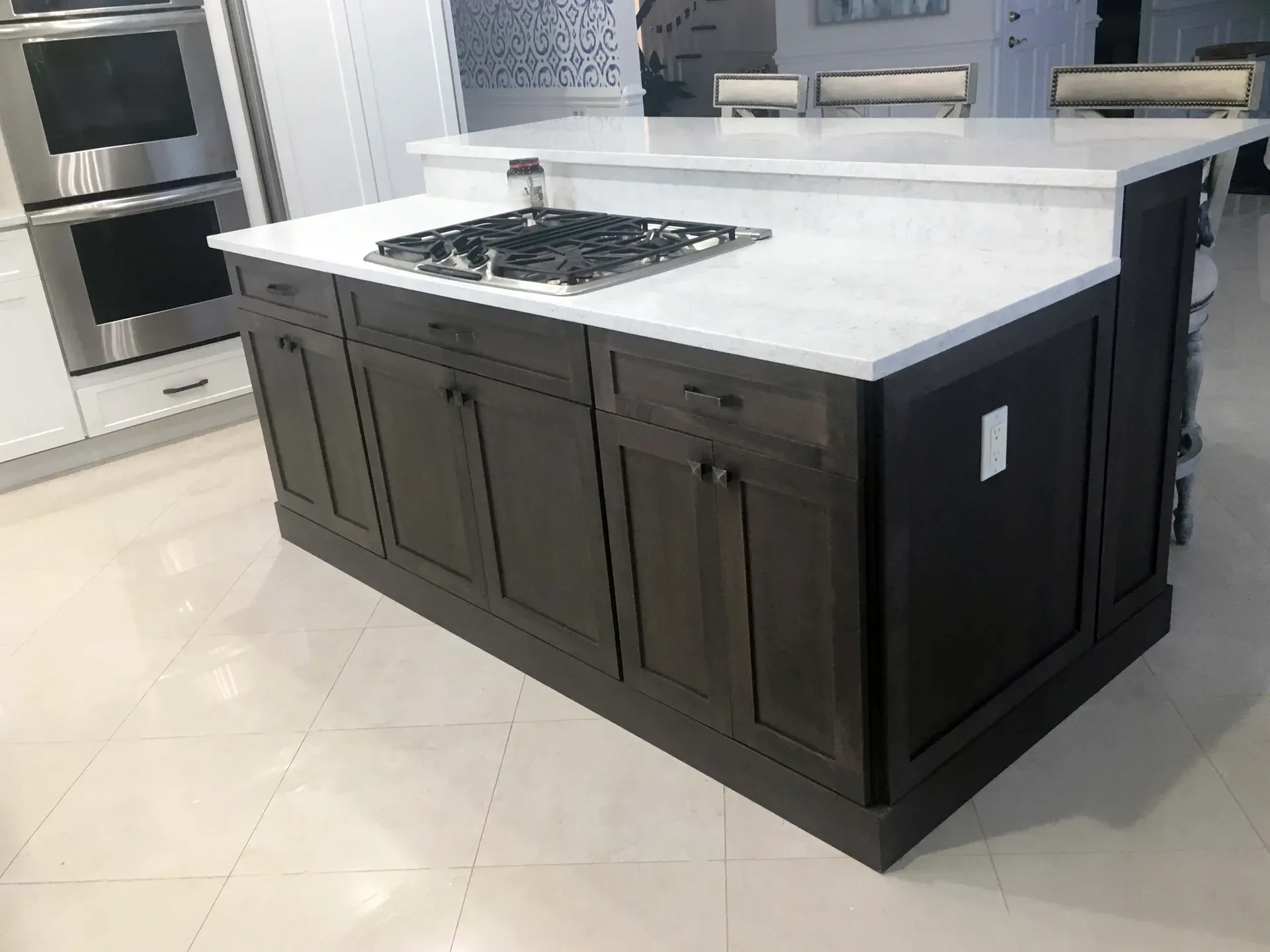A dark-stained kitchen island with a white quartz waterfall top and a gas cooktop, against white perimeter cabinets and stainless wall ovens