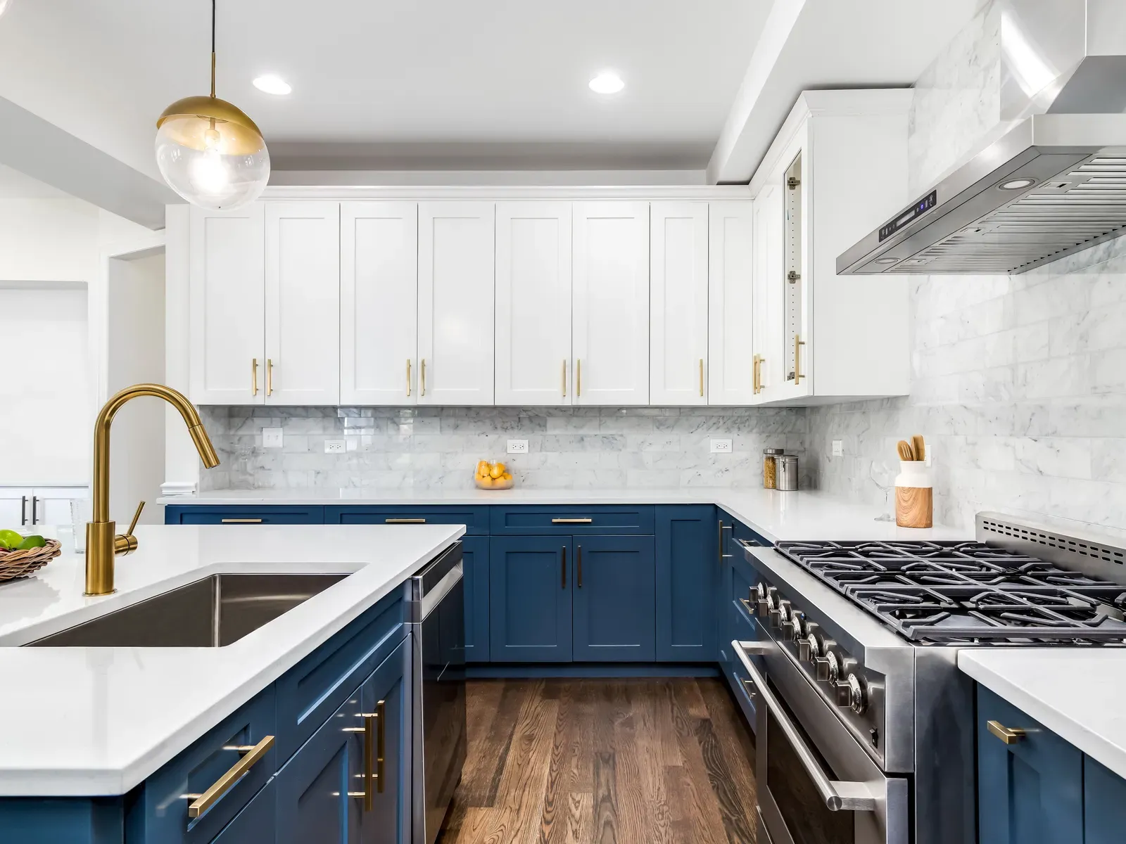 A two-tone kitchen with white shaker upper cabinets, navy base cabinets, brushed-brass hardware and a marble tile backsplash behind a stainless range