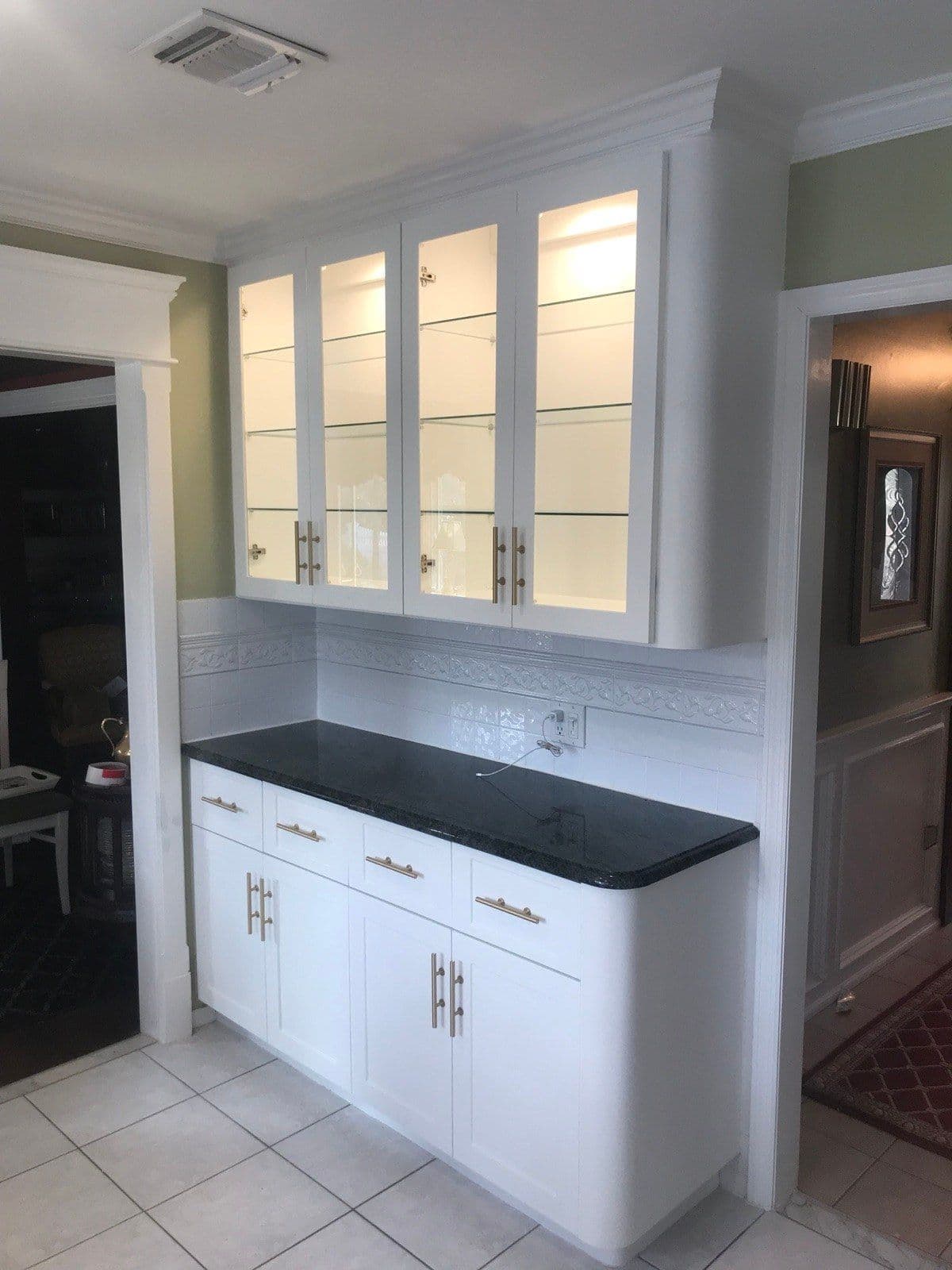 White shaker base cabinets with brushed-brass bar pulls under a black granite counter, below glass-front upper cabinets with lit glass shelves