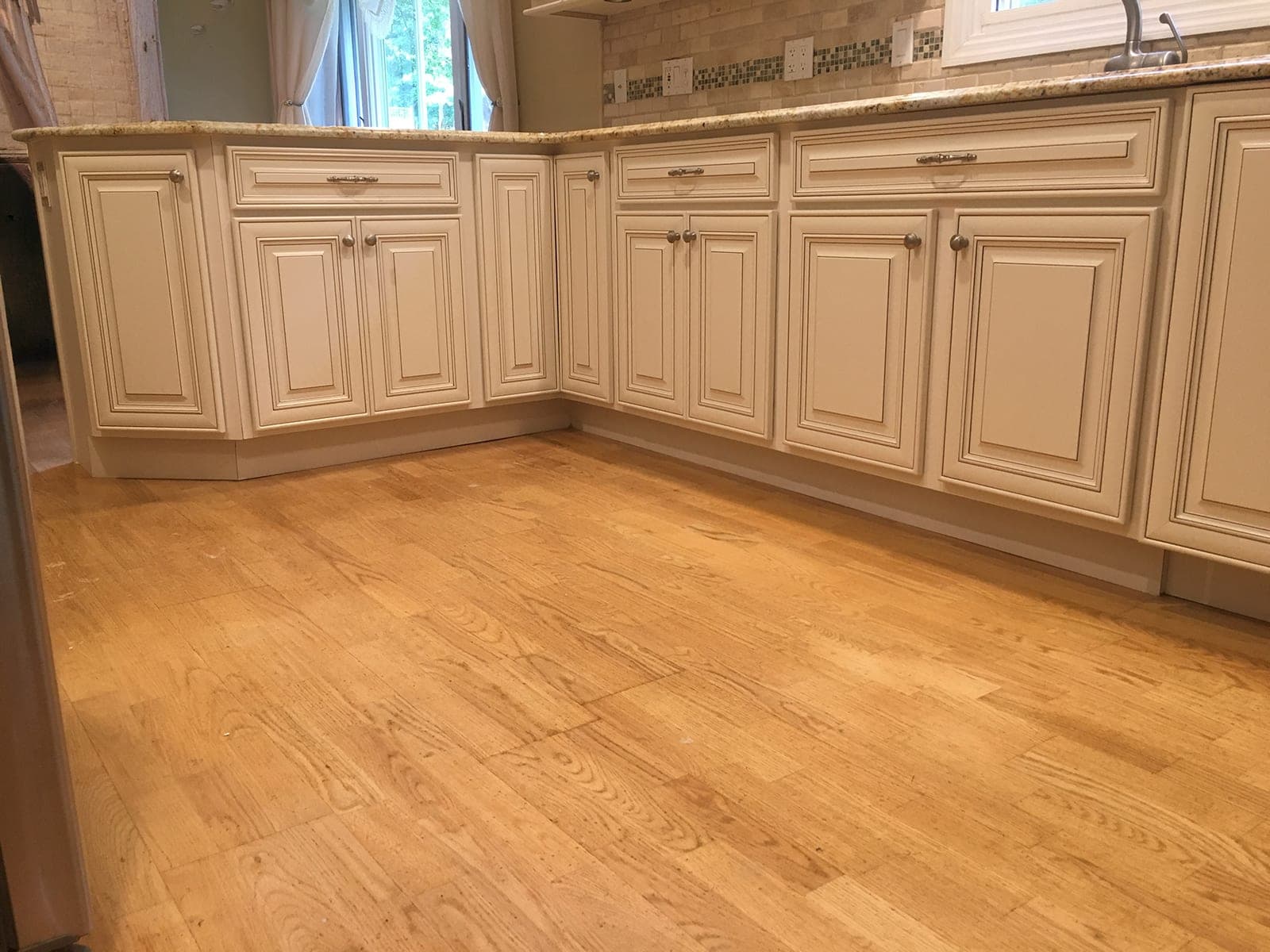 Cream raised-panel base cabinets with glazed edges and brushed-nickel knobs, wrapping an L under a gold granite counter above an oak floor