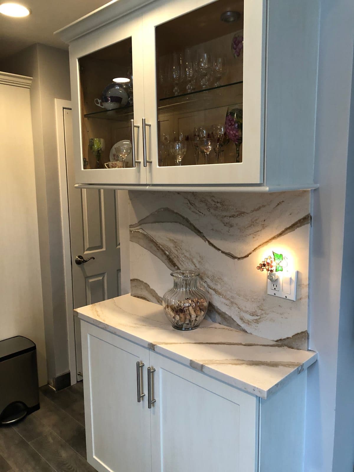 A beverage station in white shaker cabinets, with glass-front uppers and a dramatically veined stone backsplash run full height