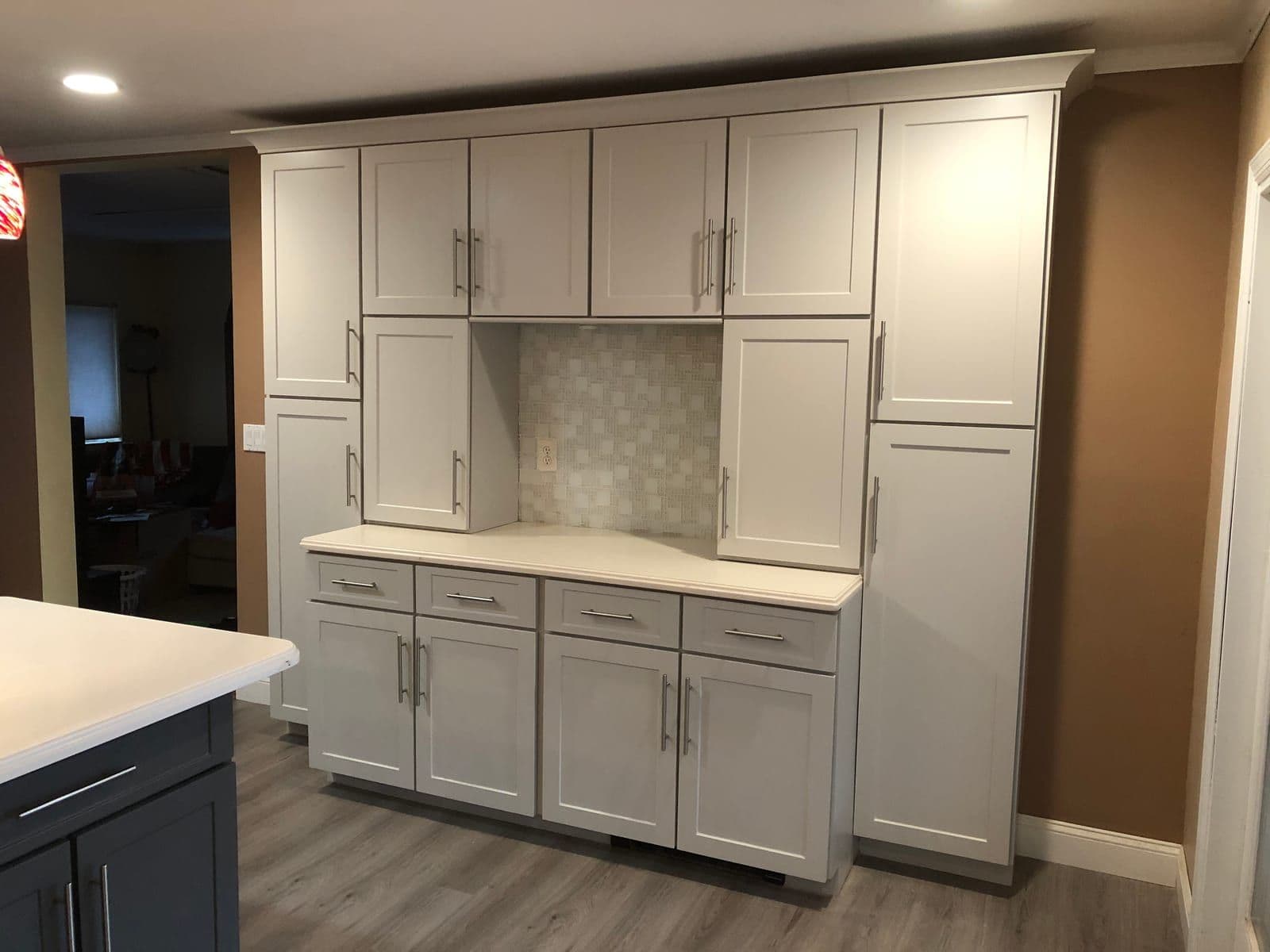 A light grey shaker pantry and coffee wall with a woven tile backsplash, beside a navy island