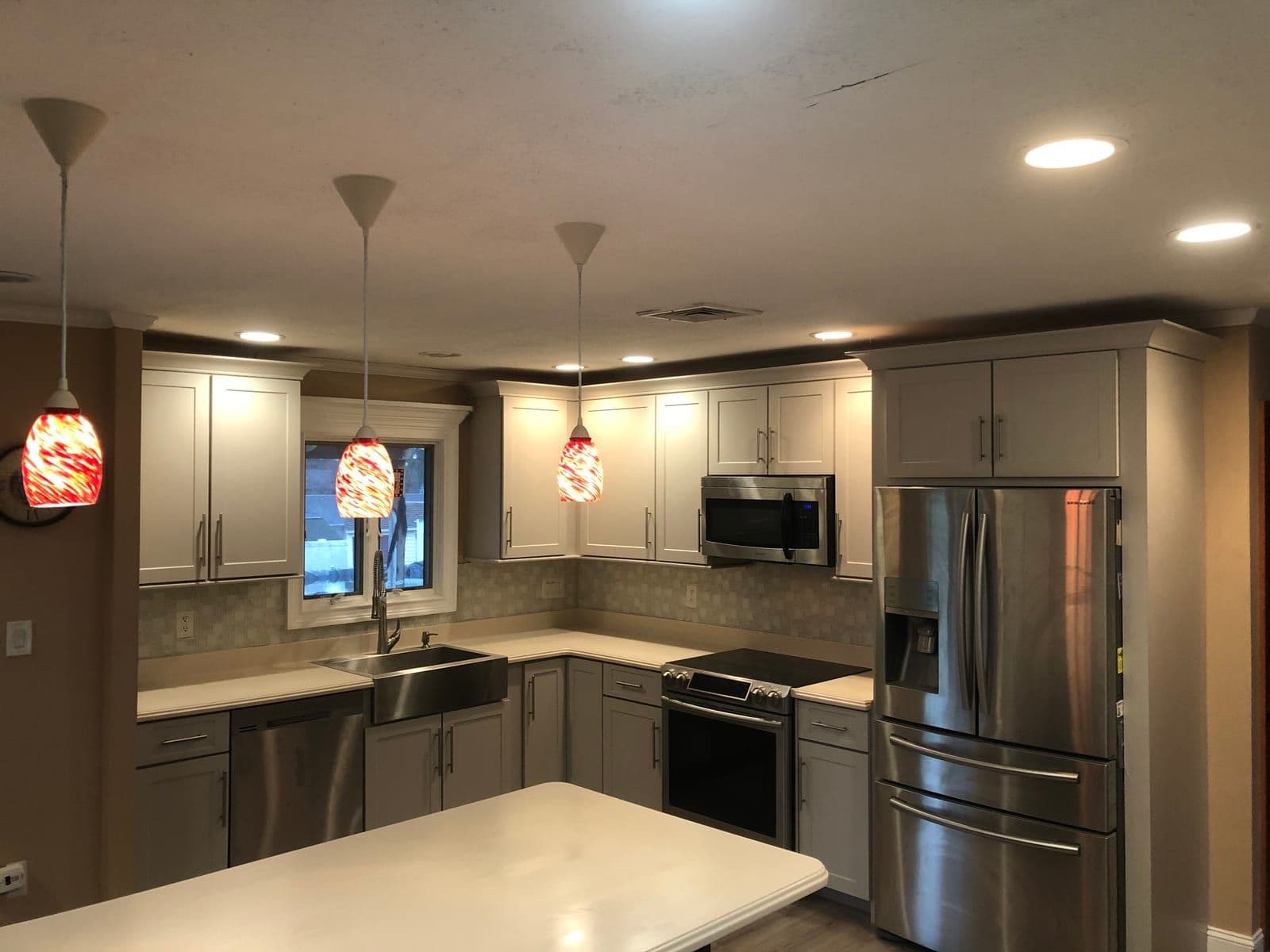 Greige shaker cabinets around a stainless farmhouse sink, under red art-glass pendants