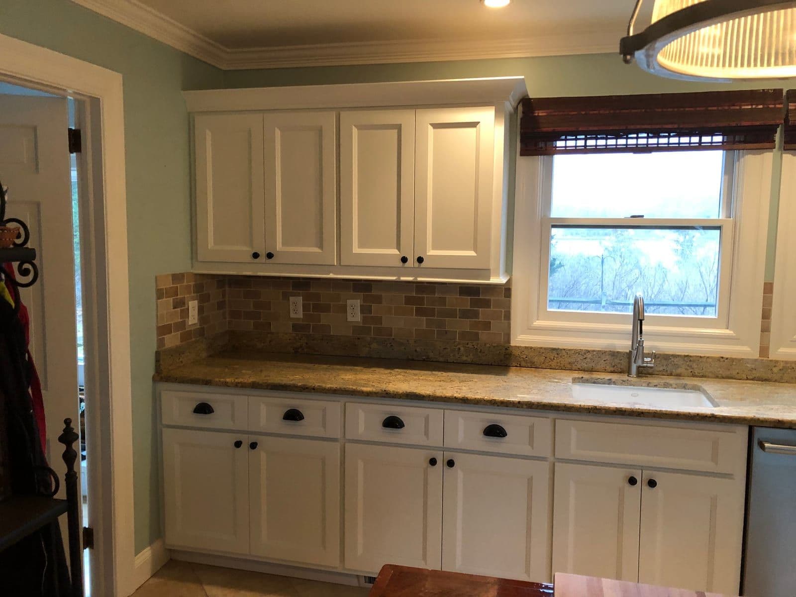 White cabinets with black cup pulls against mint-green walls, under speckled granite beside a garden window