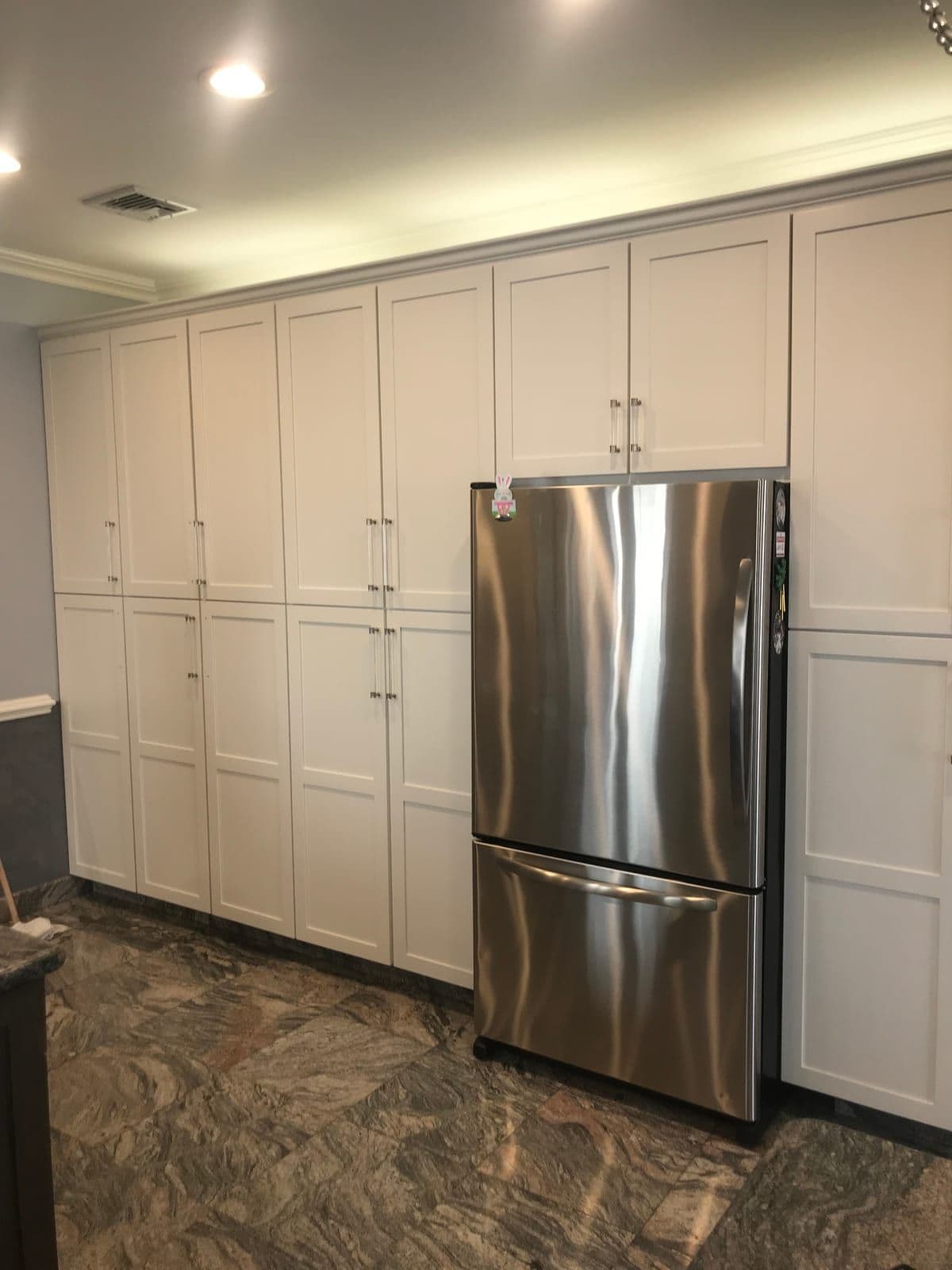 A floor-to-ceiling wall of white shaker pantry cabinets flanking a stainless refrigerator, over a veined marble floor