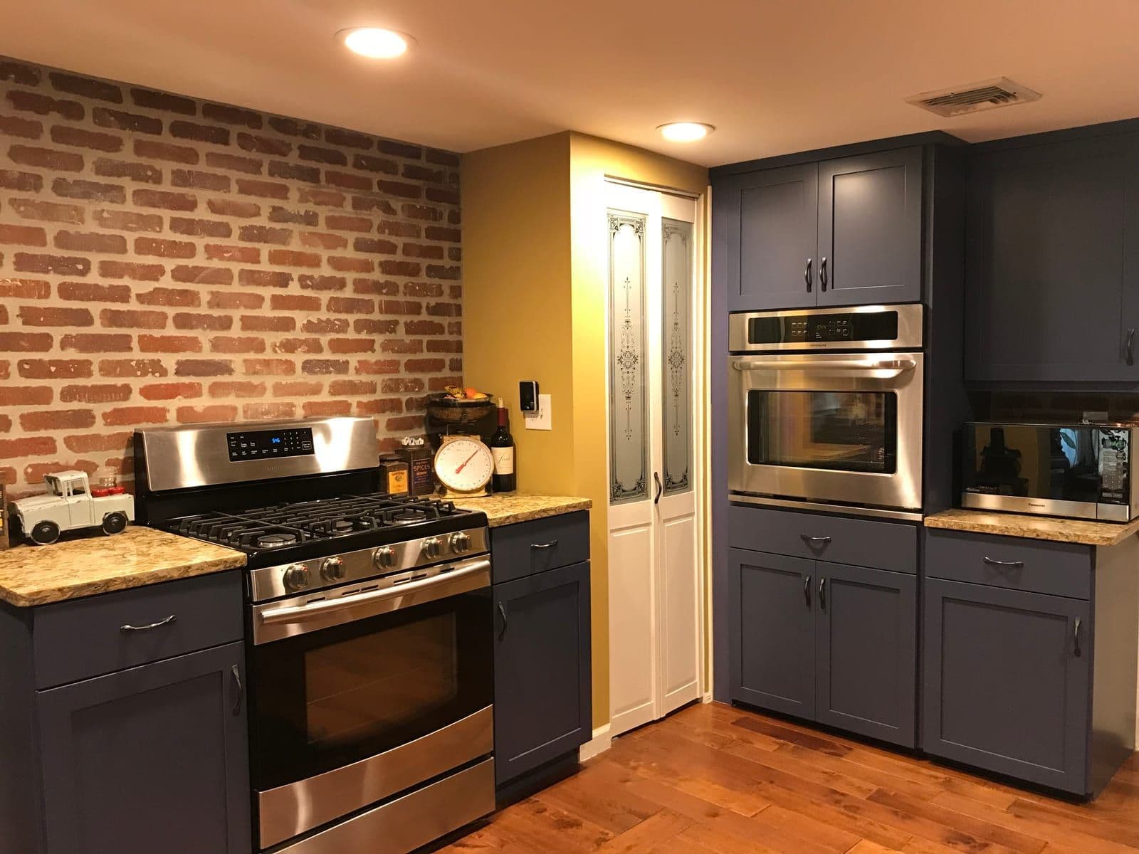 Slate-blue shaker cabinets and stainless wall ovens against an exposed red brick wall