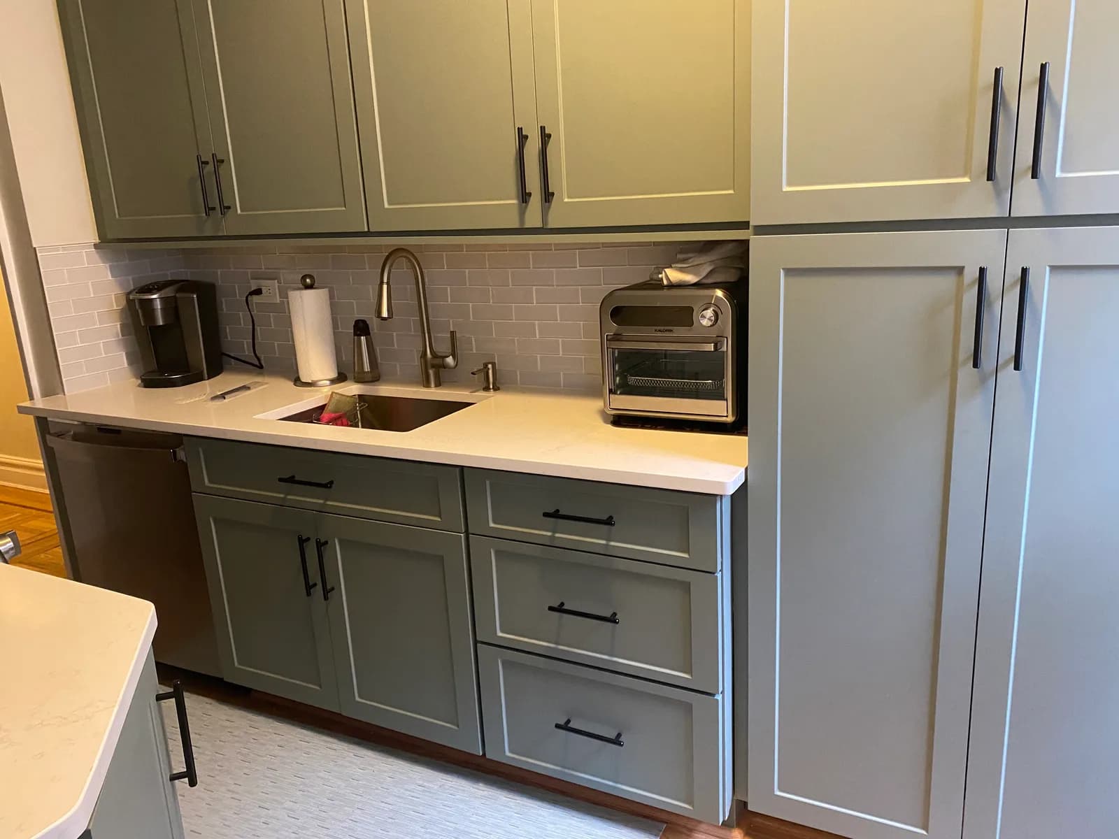A sage-green shaker kitchen with matte black bar pulls, a white quartz counter, a gray subway-tile backsplash and an undermount sink