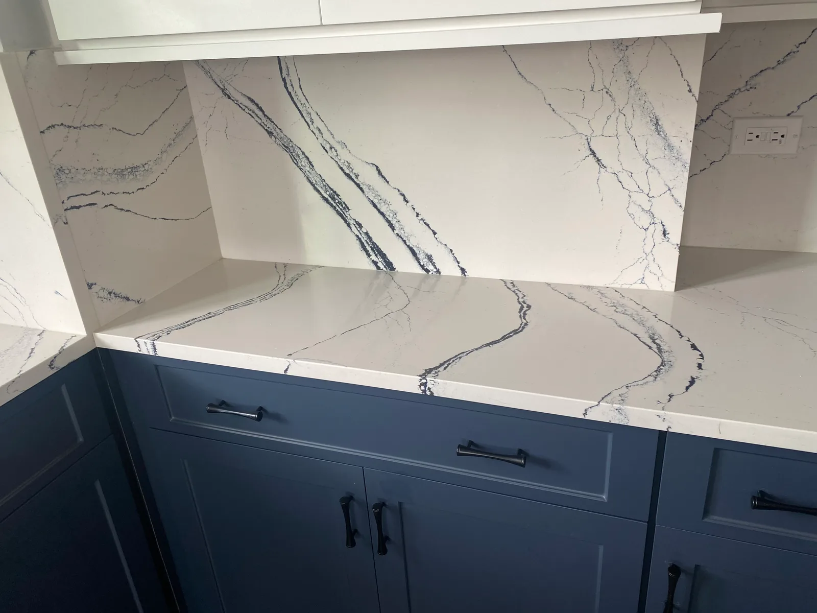 Navy shaker base cabinets with black pulls under a white quartz counter and full-height backsplash veined in dark blue