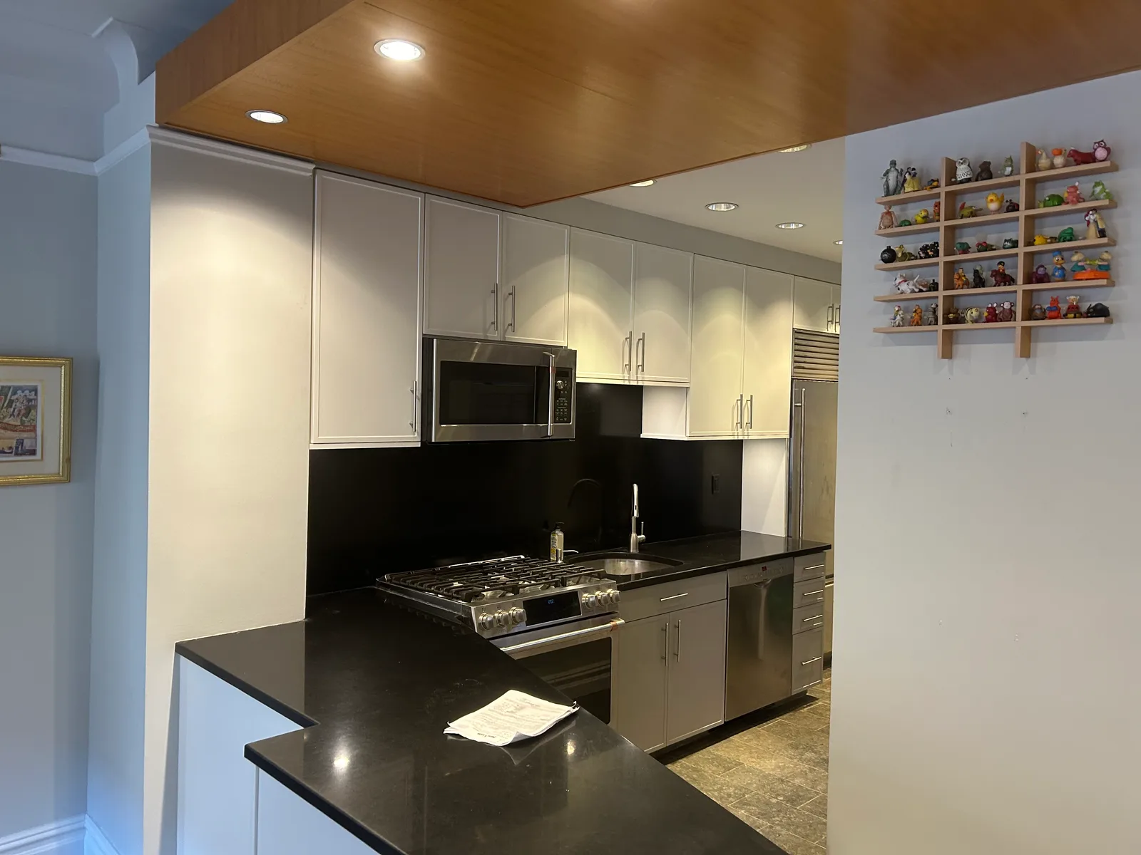 A galley-style kitchen with pale grey cabinets, a black stone counter and backsplash, a stainless gas range and a wood-paneled soffit with recessed lights