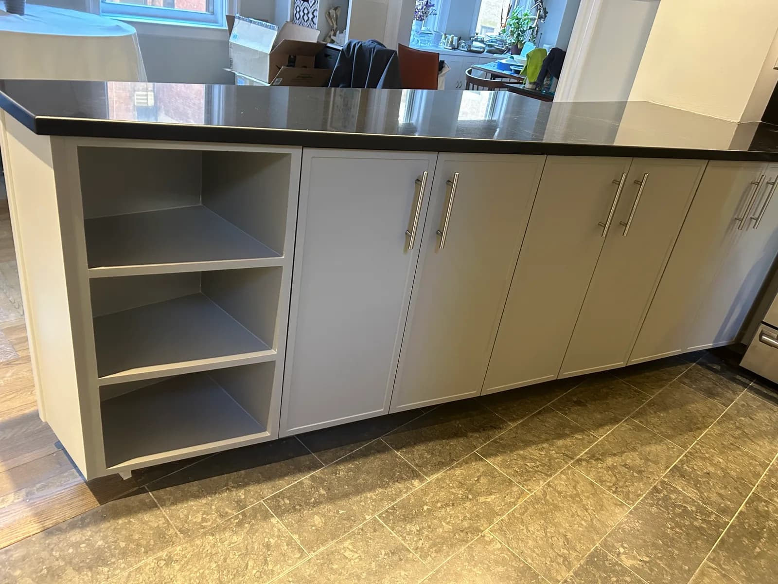 The kitchen peninsula in pale grey with brushed-steel bar pulls, three open shelves at the end and a black stone top above a stone-look tile floor