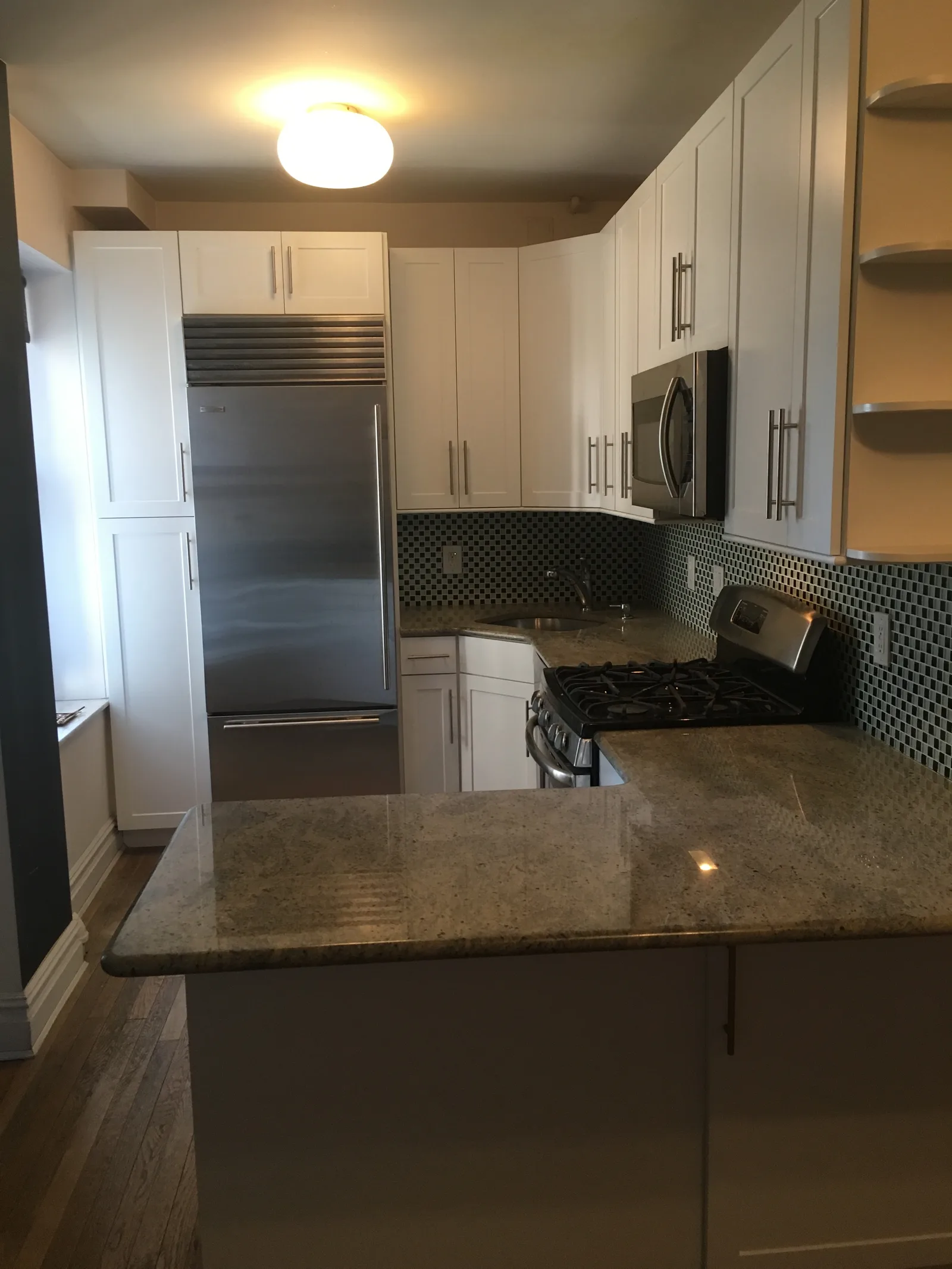 A compact white shaker kitchen with a stainless refrigerator, mosaic-tile backsplash and a granite peninsula over a wood floor