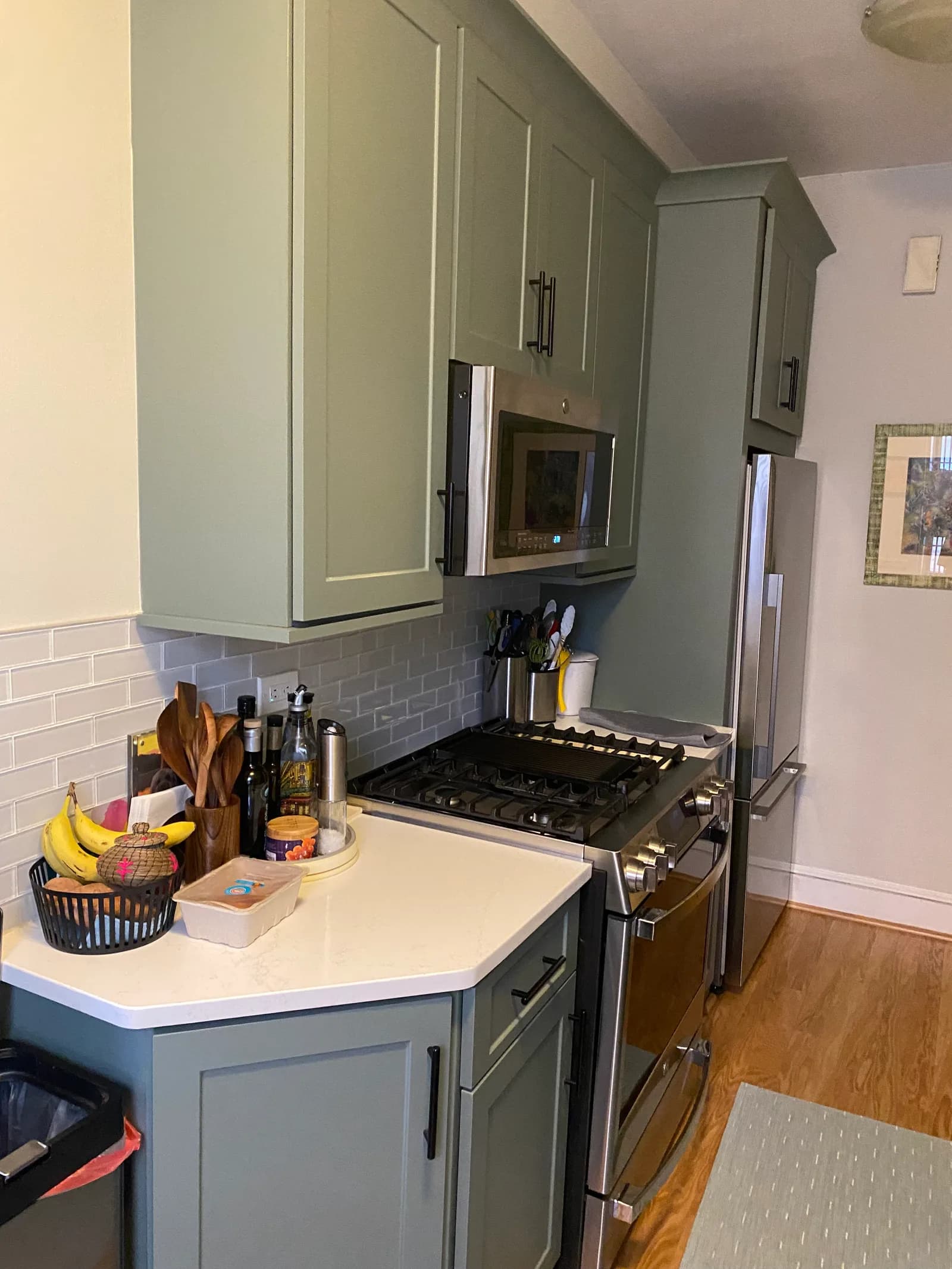 Sage-green shaker cabinets above a stainless gas range, with a white quartz counter that has a clipped corner and a gray subway-tile backsplash