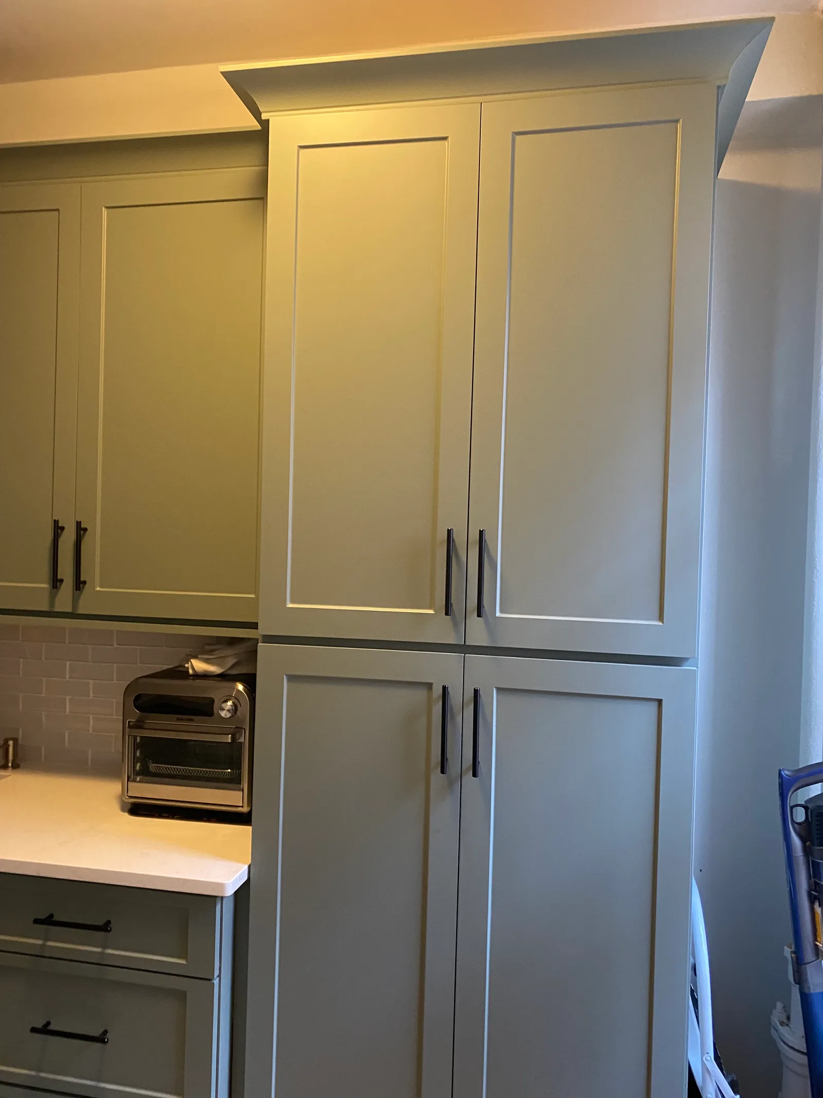 A sage-green tall pantry cabinet with crown molding and black bar pulls, beside lower drawers under a white quartz counter